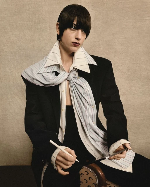 A woman with dark hair in a black blazer and a large striped tie is posing with pens in her hands in a neutral indoor setting.