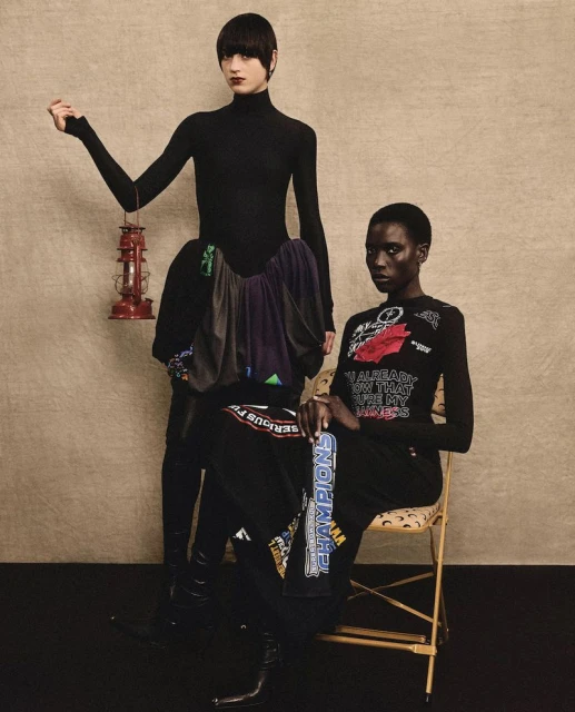 Two fashion models pose indoors against a neutral backdrop, one seated on a yellow chair and the other holding a red lantern.