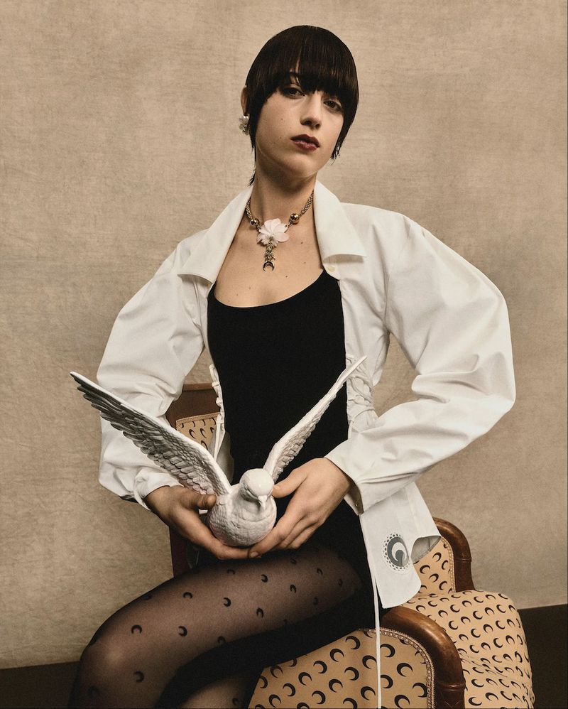 A woman sits on a patterned chair while holding a decorative white bird, wearing a black top, patterned tights, and a white jacket.