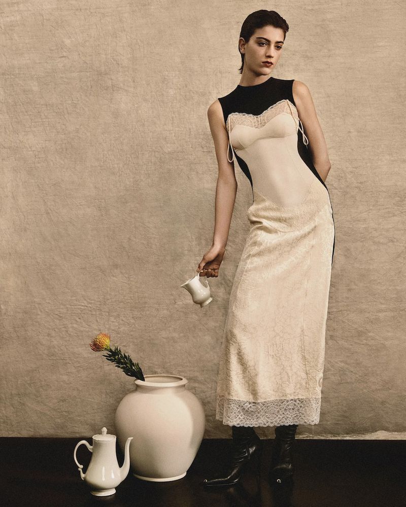 Woman in dress poses in photo studio with flower vase and teapot. Beige textured background in a fashion editorial shot.