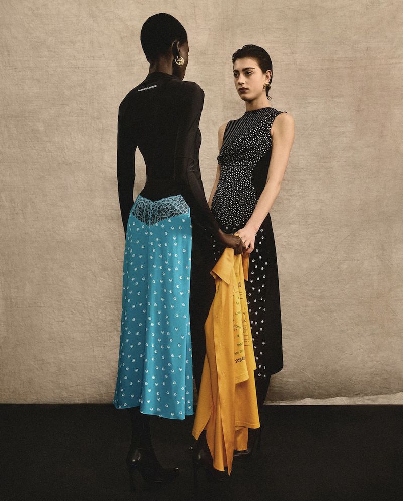 Two women in contrasting polka-dot dresses pose indoors against a neutral wall, one holding a mustard-yellow garment, fashion studio shot.