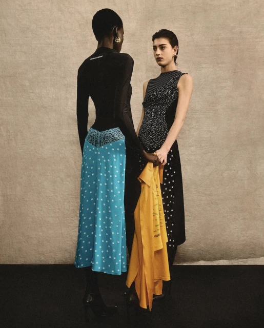 Two women in contrasting polka-dot dresses pose indoors against a neutral wall, one holding a mustard-yellow garment, fashion studio shot.