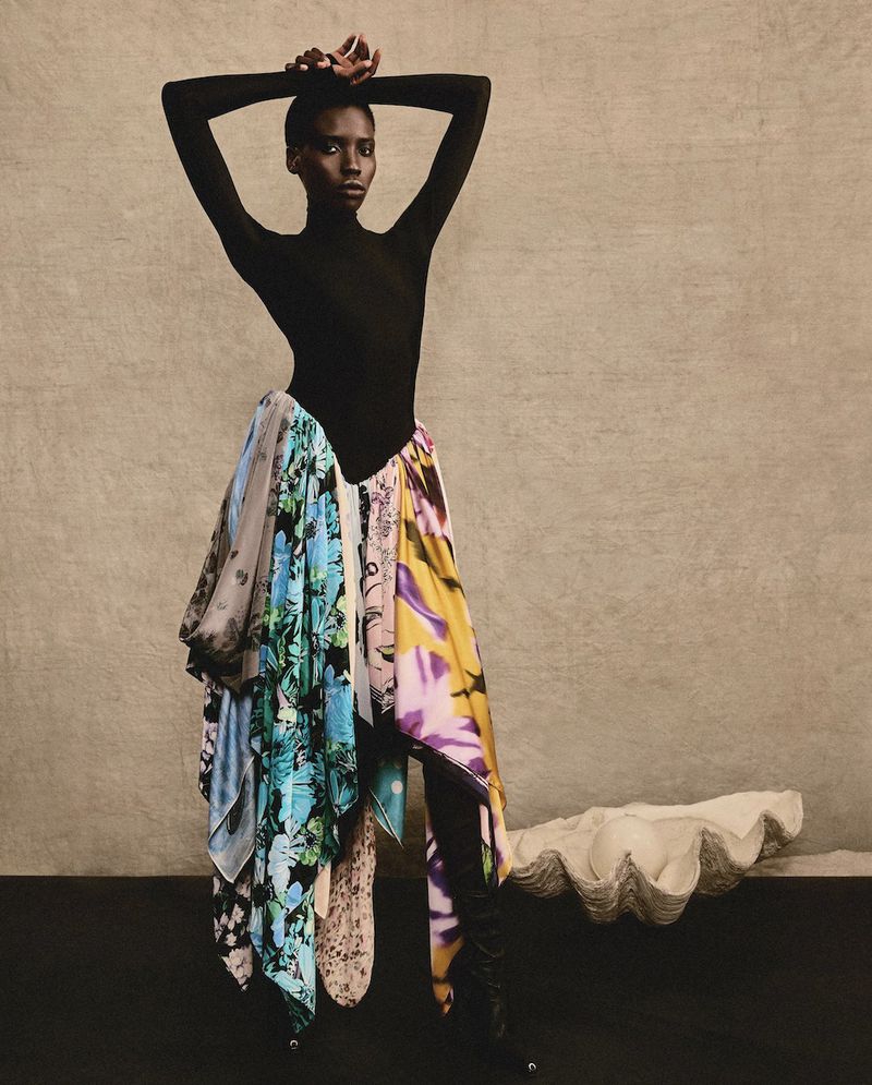 Fashionable woman poses in black bodysuit and scarf skirt against neutral backdrop. Studio shot, portrait, full-length.