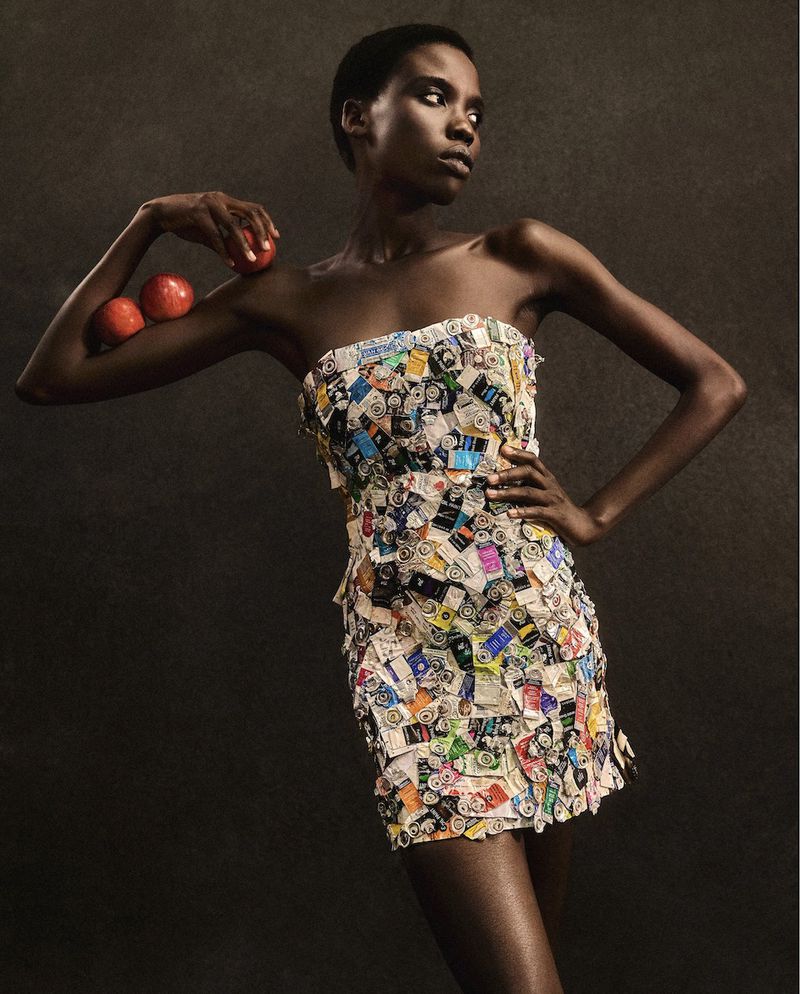 An African-American woman is posing in a mini dress made of assorted pictures and holding red apples in front of a dark background.