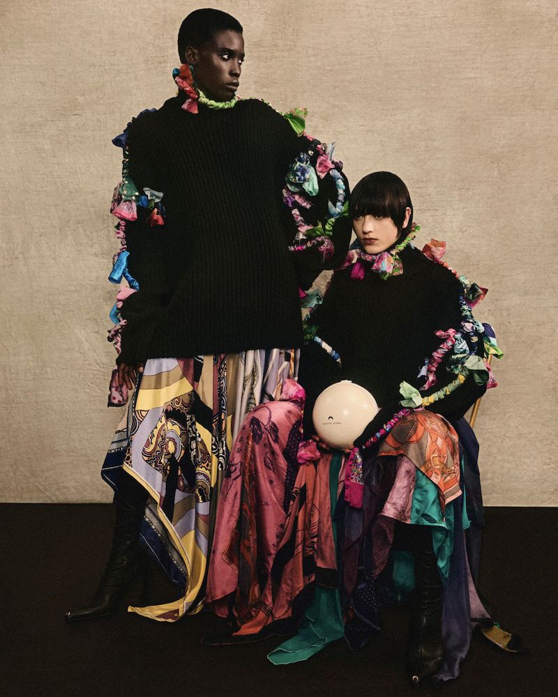 Two fashion models pose in black sweaters and flowing patterned skirts, accessorized with colorful ruffled embellishments, against neutral backdrop.