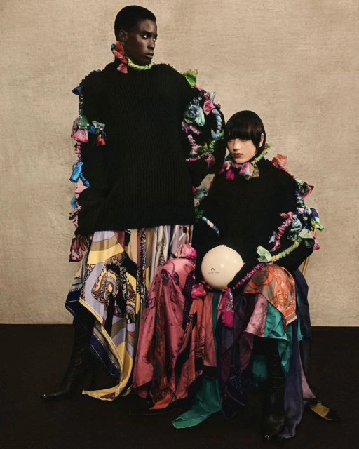 Two fashion models pose in black sweaters and flowing patterned skirts, accessorized with colorful ruffled embellishments, against neutral backdrop.