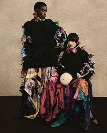 Two fashion models pose in black sweaters and flowing patterned skirts, accessorized with colorful ruffled embellishments, against neutral backdrop.