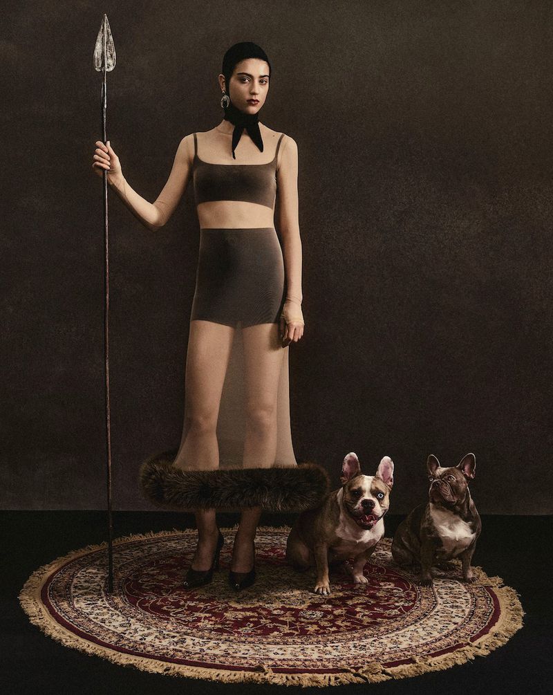 Woman in modern ensemble poses with spear and French bulldogs on oriental rug in a studio, creating a unique portrait.