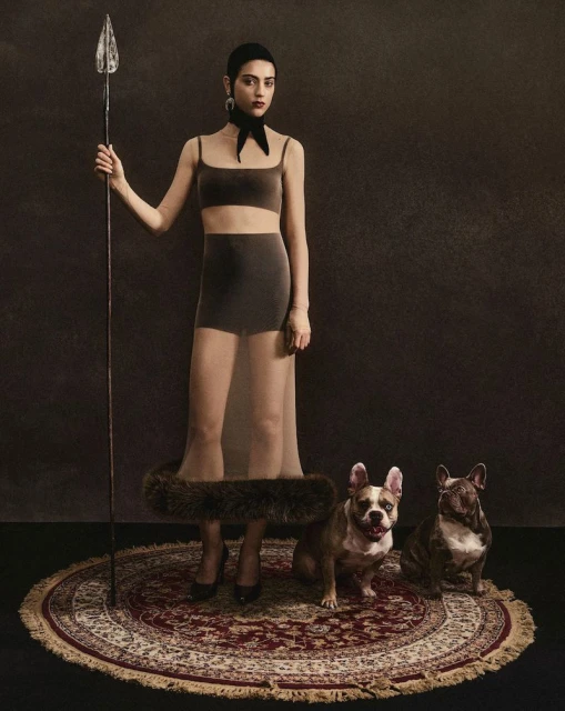 Woman in modern ensemble poses with spear and French bulldogs on oriental rug in a studio, creating a unique portrait.