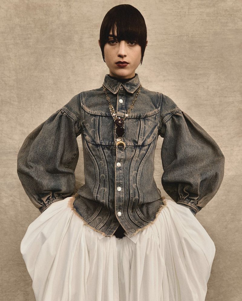 A woman with dark hair and bangs poses in a studio wearing a unique denim and white dress with statement sleeves and a gold pendant necklace.