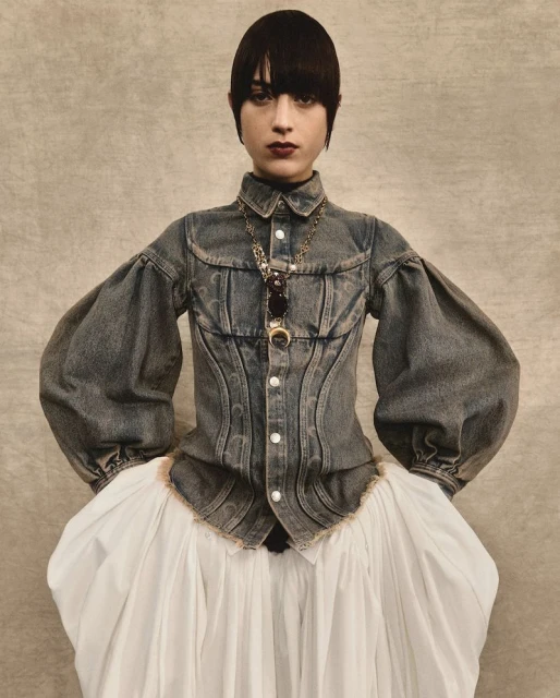 A woman with dark hair and bangs poses in a studio wearing a unique denim and white dress with statement sleeves and a gold pendant necklace.