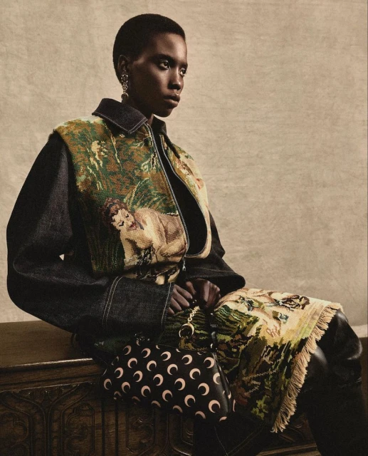 Dark-skinned woman sits on a wooden chest wearing a jacket with nature tapestry, holding a black crescent moon print purse.