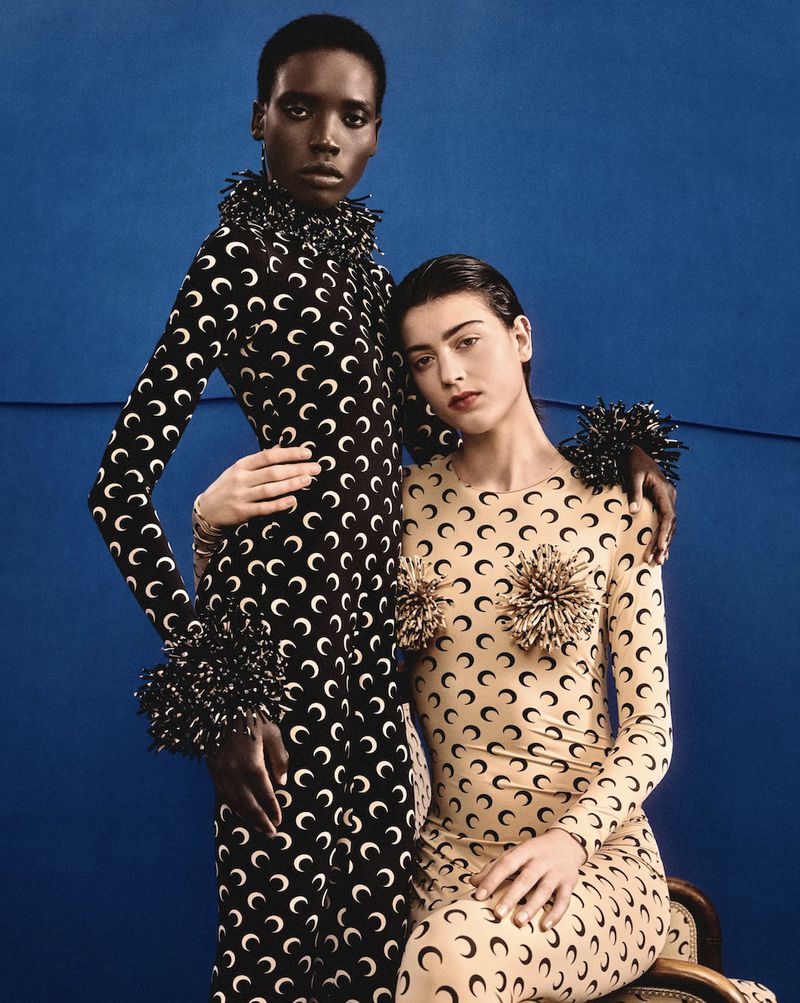 Two women pose in matching long-sleeved dresses with a moon crescent pattern against a blue backdrop in a fashion editorial shot.