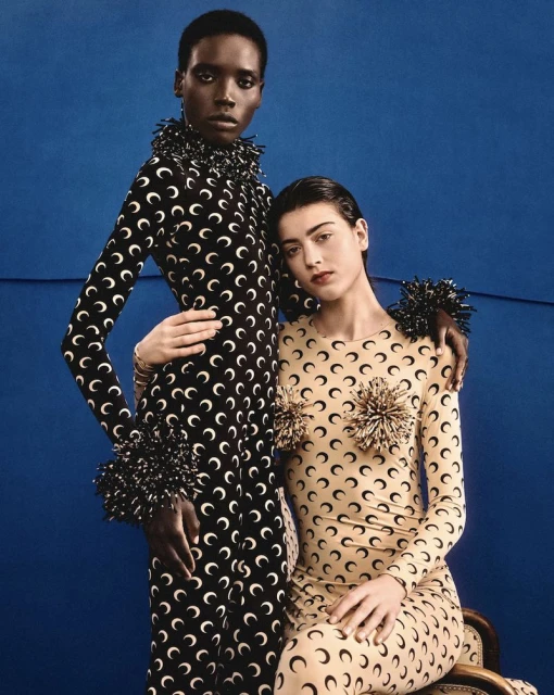 Two women pose in matching long-sleeved dresses with a moon crescent pattern against a blue backdrop in a fashion editorial shot.