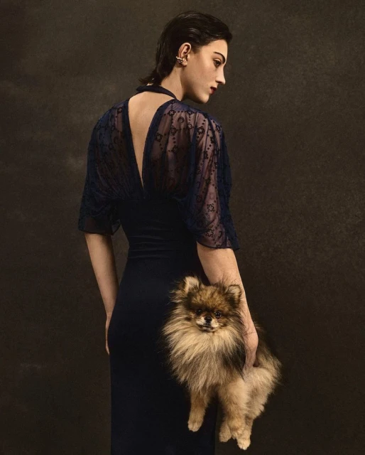Woman in navy dress posing with a small dog. The dog is in focus. Dark background. Low-key lighting. Fashion photography.