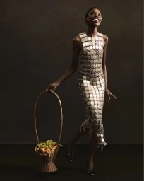 Model in a metallic disk dress holding a tall wicker basket of fruit against a dark backdrop for Marine Serre Fall 2026