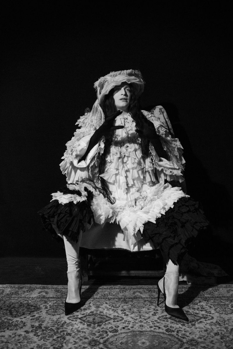 Monochrome portrait of a person wearing a frilled costume with black gloves and high heels, seated on a chair on a patterned carpet.