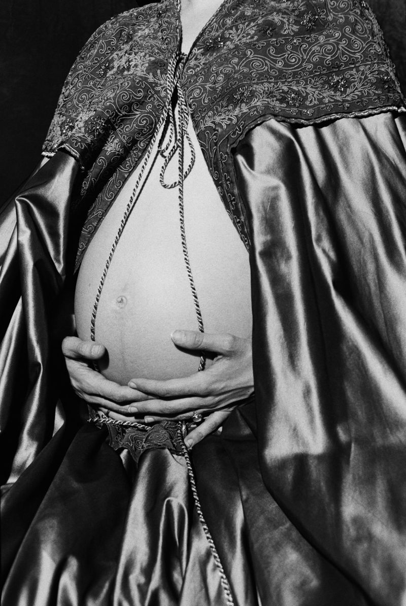 Monochromatic shot of a pregnant person with hands on their stomach, wearing a decorative cape, photographed indoors with soft light.