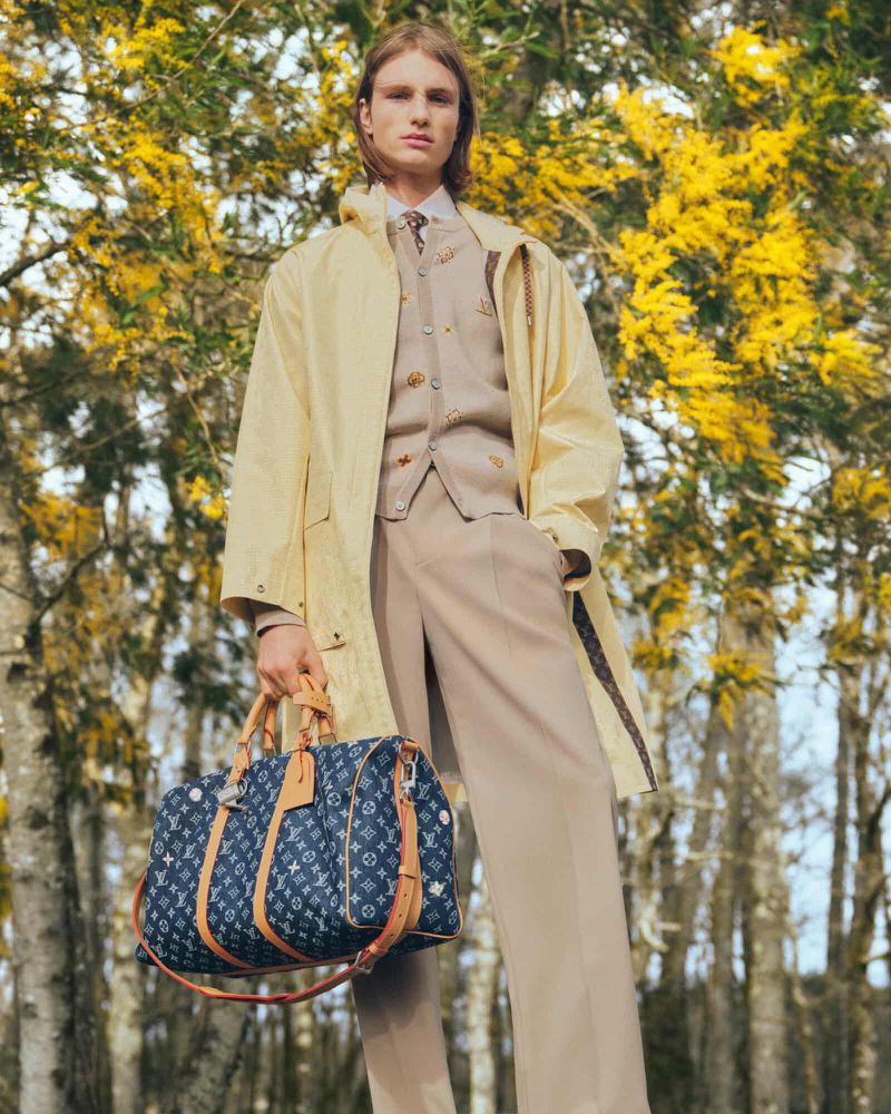 A fair-skinned male model poses in a beige suit and light yellow jacket with a Louis Vuitton bag in a sunny forest setting.