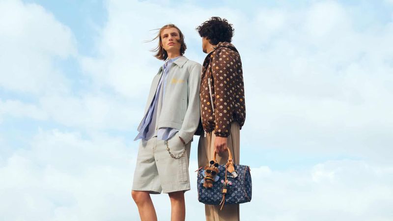 Two fashion models pose with a Louis Vuitton bag against a blue sky with light clouds, showcasing stylish jackets and shorts.