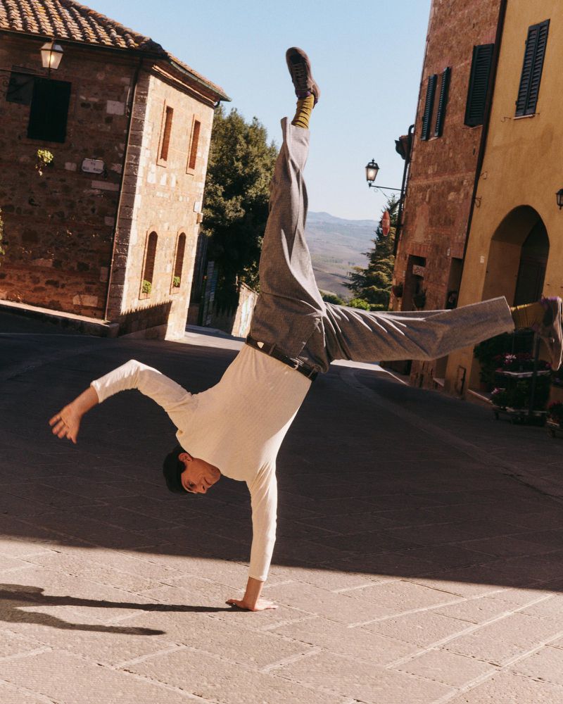 Adult male doing a handstand on a brick street in a European village with beige buildings and a clear sky on a sunny day.