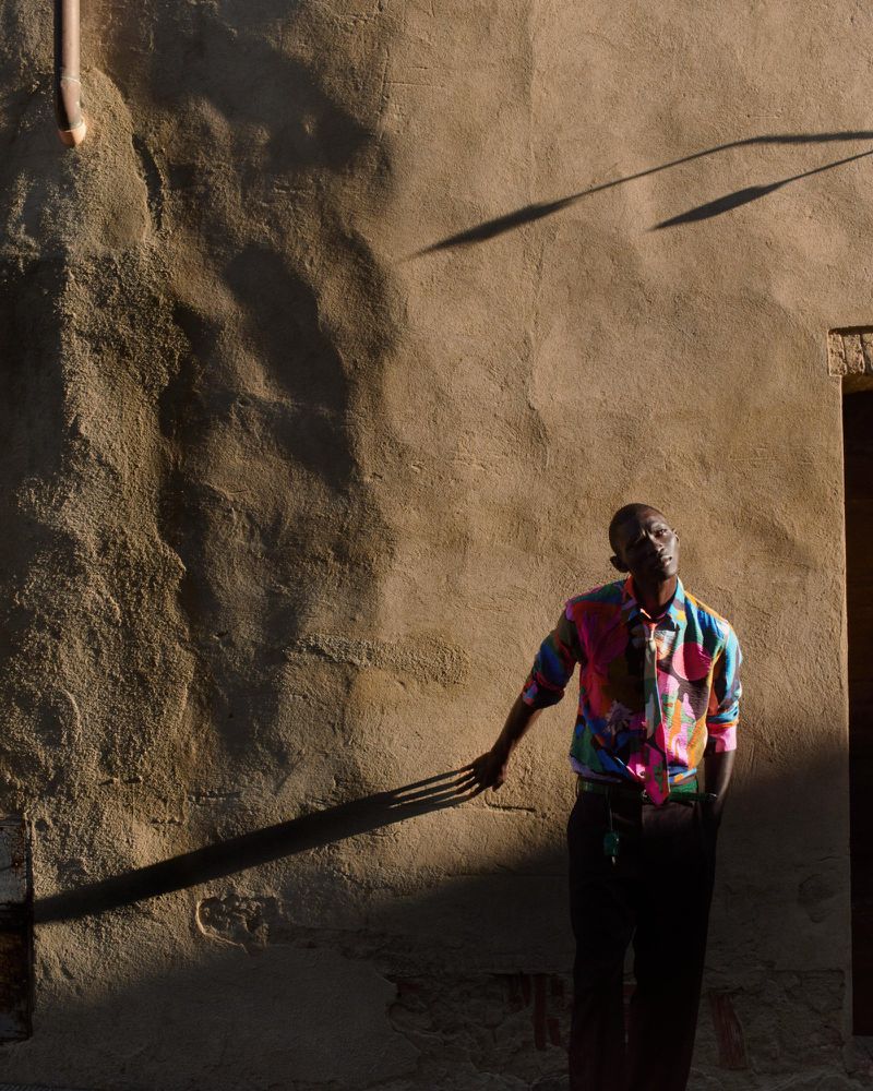 Portrait of an adult male in brightly colored shirt against weathered wall in dappled sunlight, wearing dark trousers and tie.