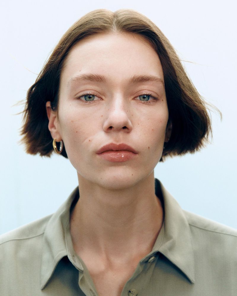 Close-up portrait of a model with short brown hair wearing a muted green collared shirt against a plain white background.