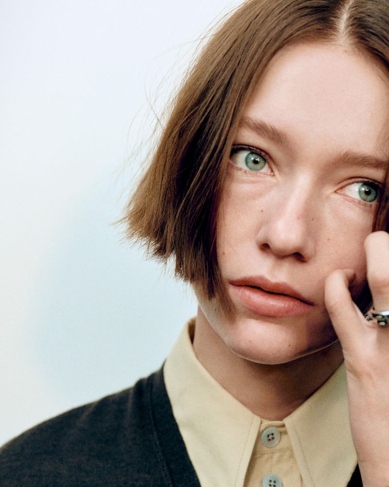 Close-up portrait of a young adult female with short brown hair, pale skin, and light green eyes, wearing a yellow shirt and grey sweater.