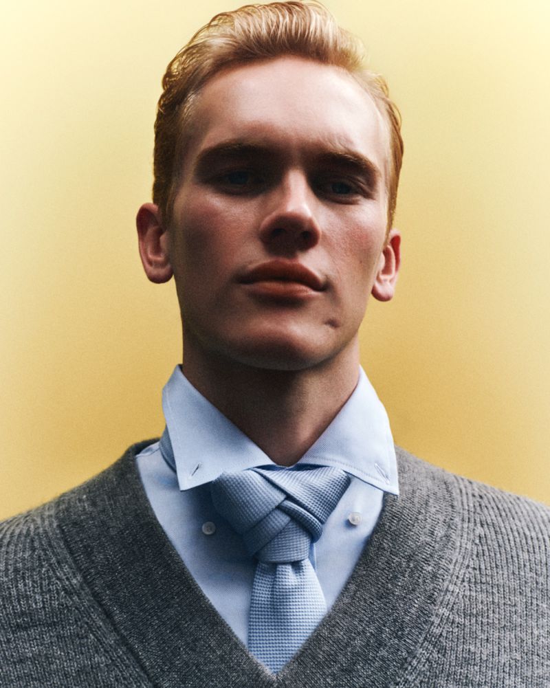 Close-up portrait of a fair-skinned young adult male with blond hair wearing a sweater and knotted tie against a yellow background.