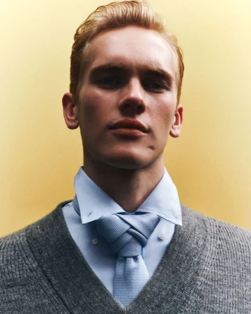 Close-up portrait of a fair-skinned young adult male with blond hair wearing a sweater and knotted tie against a yellow background.