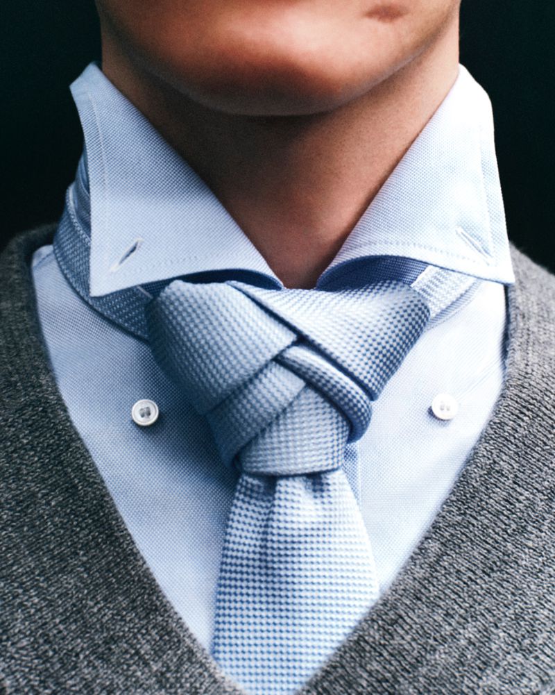 Close-up of a man's shirt and tie, with the tie knotted in an unusual way, paired with a grey sweater. Focus on textures and style.