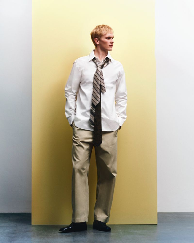 Man in white shirt, beige pants, and tie poses against a two-tone yellow and white background, looking off to the side with hands in pockets.