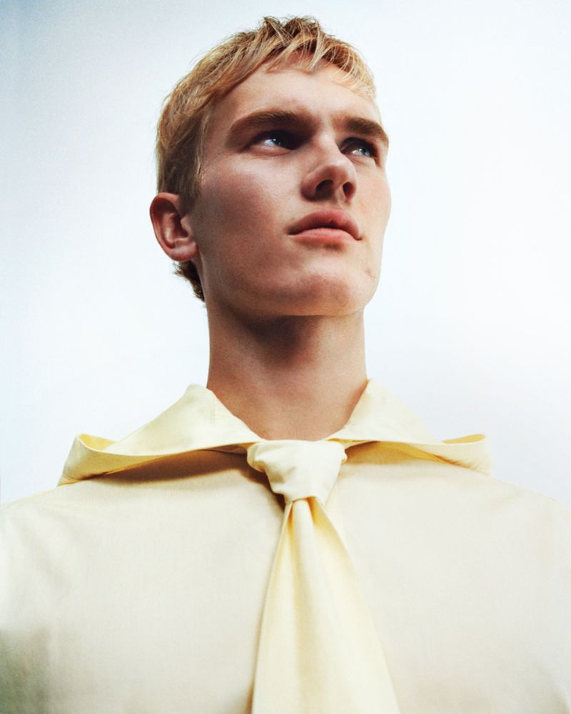 Portrait of a young adult male with blond hair looking up, wearing a light yellow shirt and tie against a bright, neutral background.