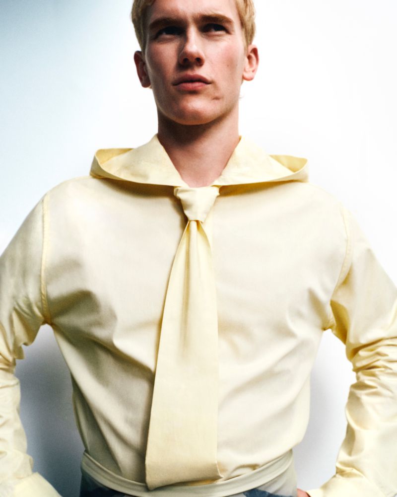Studio portrait of a blond male model wearing a pale yellow shirt with a wide collar, tied at the neck, against a white background.