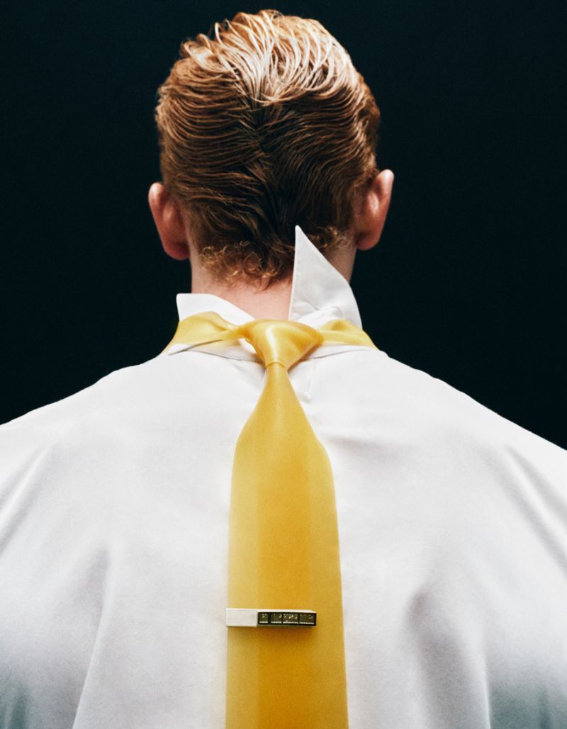 Back view of an adult male with light-colored hair wearing a white collared shirt and a yellow tie with a tie clip against black background.