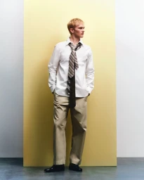 Blonde male model wearing white shirt, striped tie and beige trousers standing against a yellow and white studio backdrop