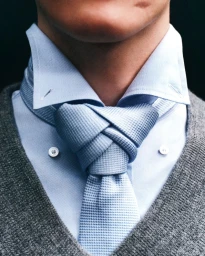 Close-up portrait of a man wearing a light blue dress shirt, textured light blue tie, and grey V-neck sweater.