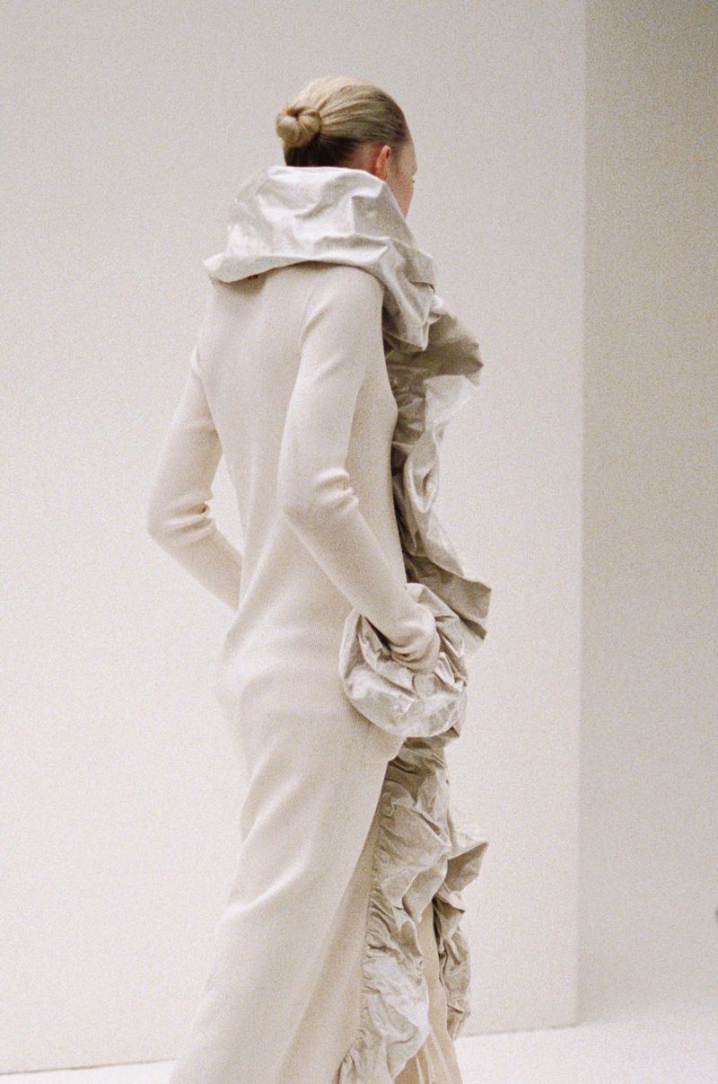 Back view of a blonde female model in a light beige, long-sleeved dress with large ruffles, posing against a plain white backdrop in a fashion setting.