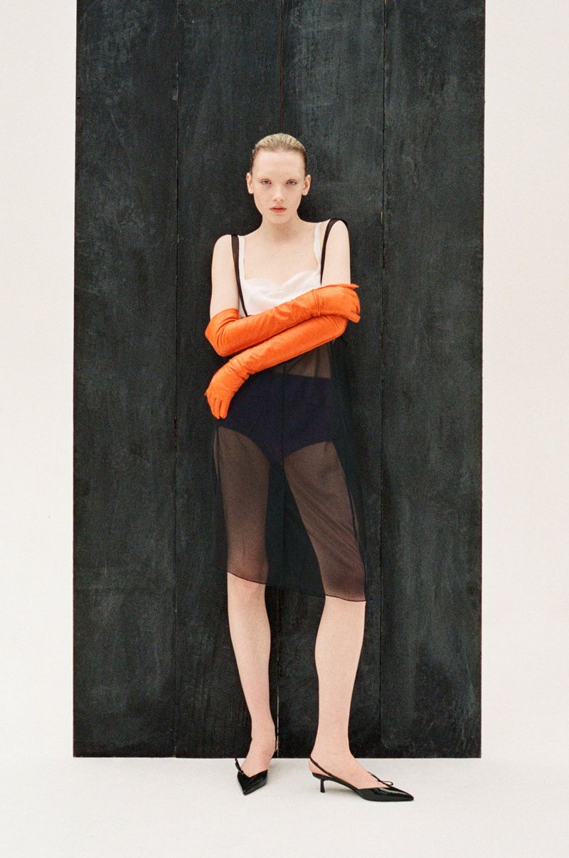 Fashion photo of a woman in a sleeveless top, dark bottoms, and orange gloves, posing against a dark wood panel and a white background.
