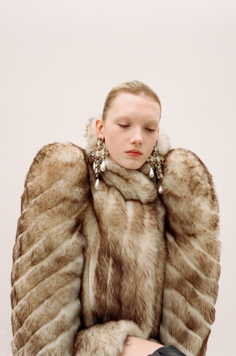 A woman with her hair pulled back wears a large, light brown faux fur coat with prominent shoulders and ornate drop earrings with faux pearls.