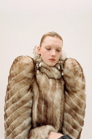 A woman with her hair pulled back wears a large, light brown faux fur coat with prominent shoulders and ornate drop earrings with faux pearls.