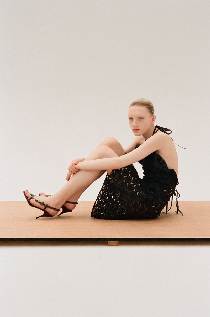 An adult woman sits on a wooden surface wearing a black dress and strappy sandals in a studio setting with a neutral background.