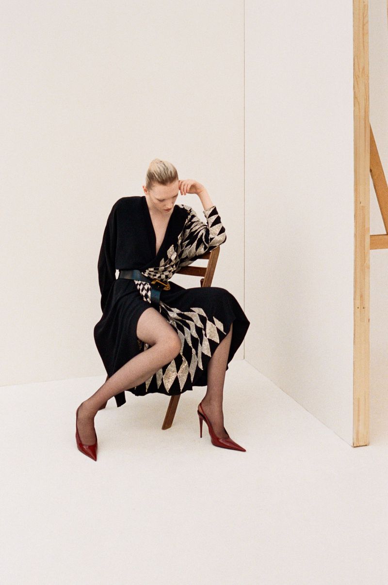 Model seated on a wooden chair wearing a black and patterned dress with red heels against a white studio wall.