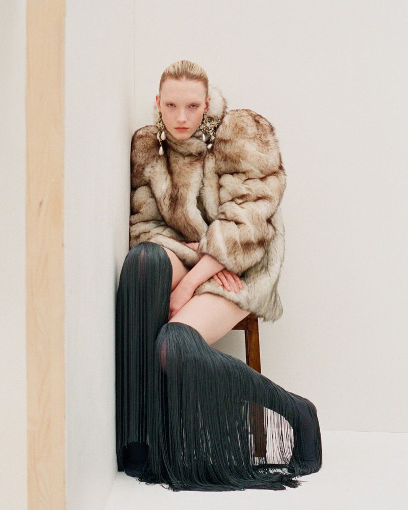 Fashion portrait of a woman in a fur coat and fringe boots sitting in a corner, looking at the camera with a neutral expression.