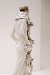 Model wearing a long white knit dress with ruffled collar standing against a clean white wall