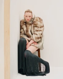 Model wearing a faux fur coat and fringe boots seated against a white wall for Muse Magazine