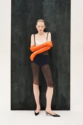 Model in a black sheer dress and long orange gloves leaning against a dark textured wooden panel