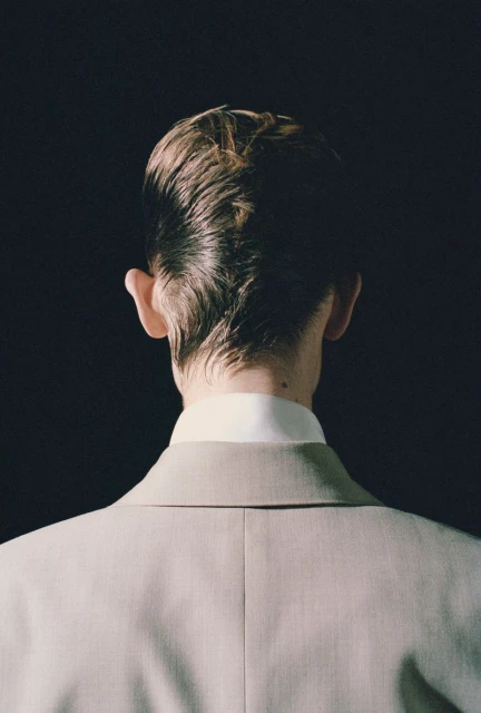 Back view of a person with combed-back blonde hair wearing a light beige suit against a dark background in a portrait orientation.