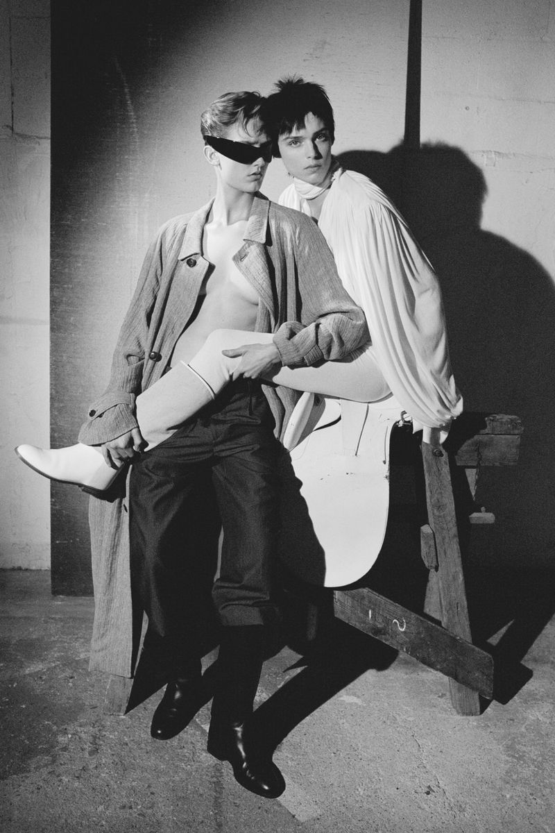 Monochrome fashion shot of two slender men in a studio, one seated on a wooden structure, the other standing, both in stylish attire.