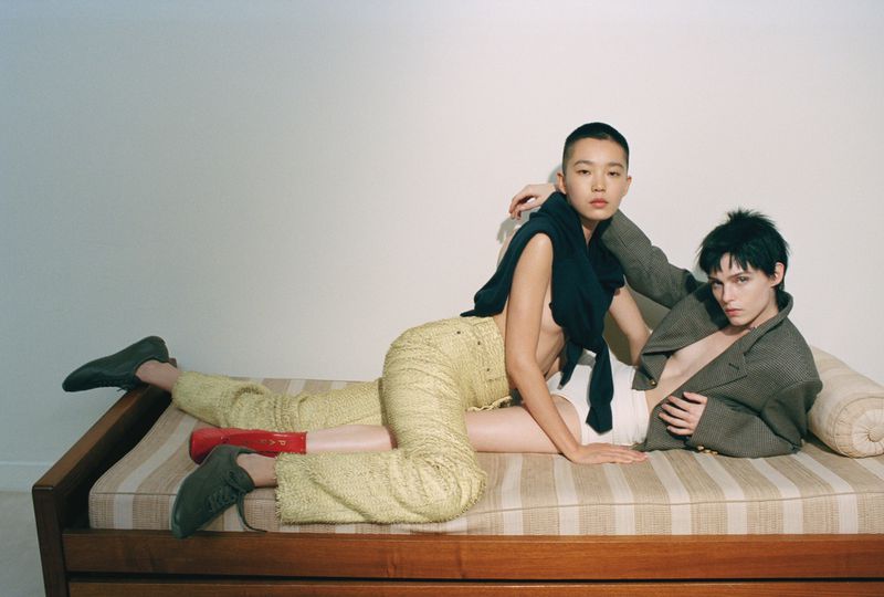 Two women in casual outfits pose intimately on a striped daybed in a brightly lit studio setting, conveying modern style and fashion.